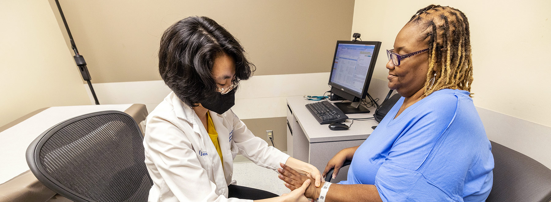 While both are seated and facing each other, Dr. Cathy Lee Ching examines a patient's hand in an exam room.